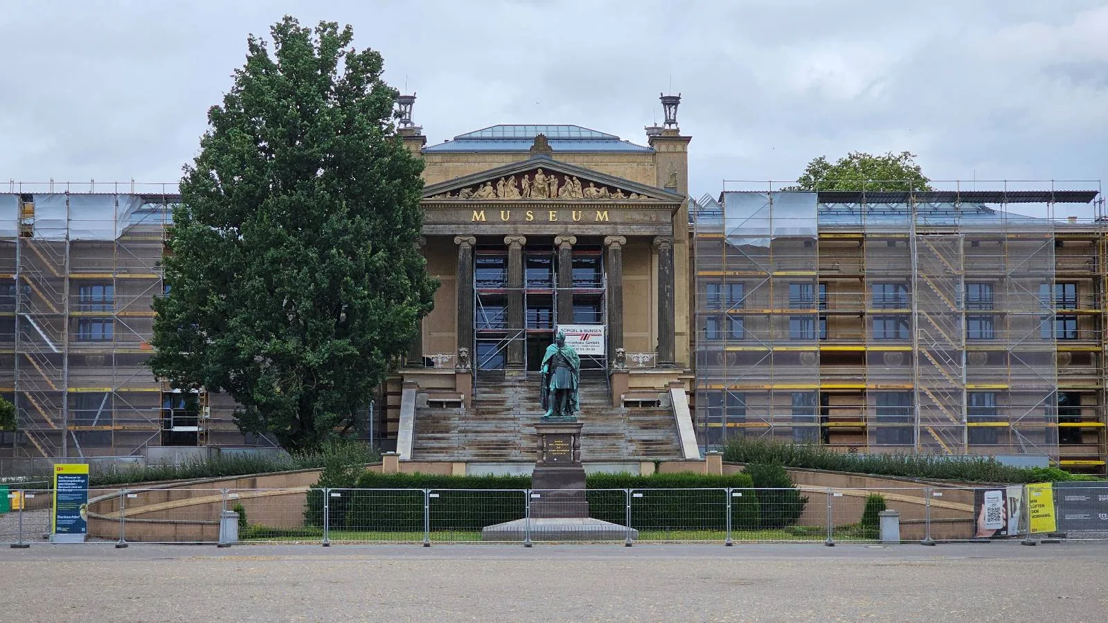 A grand neoclassical building with columns and a central staircase, partially covered in scaffolding for renovations, with a large tree on the left and cloudy sky above.