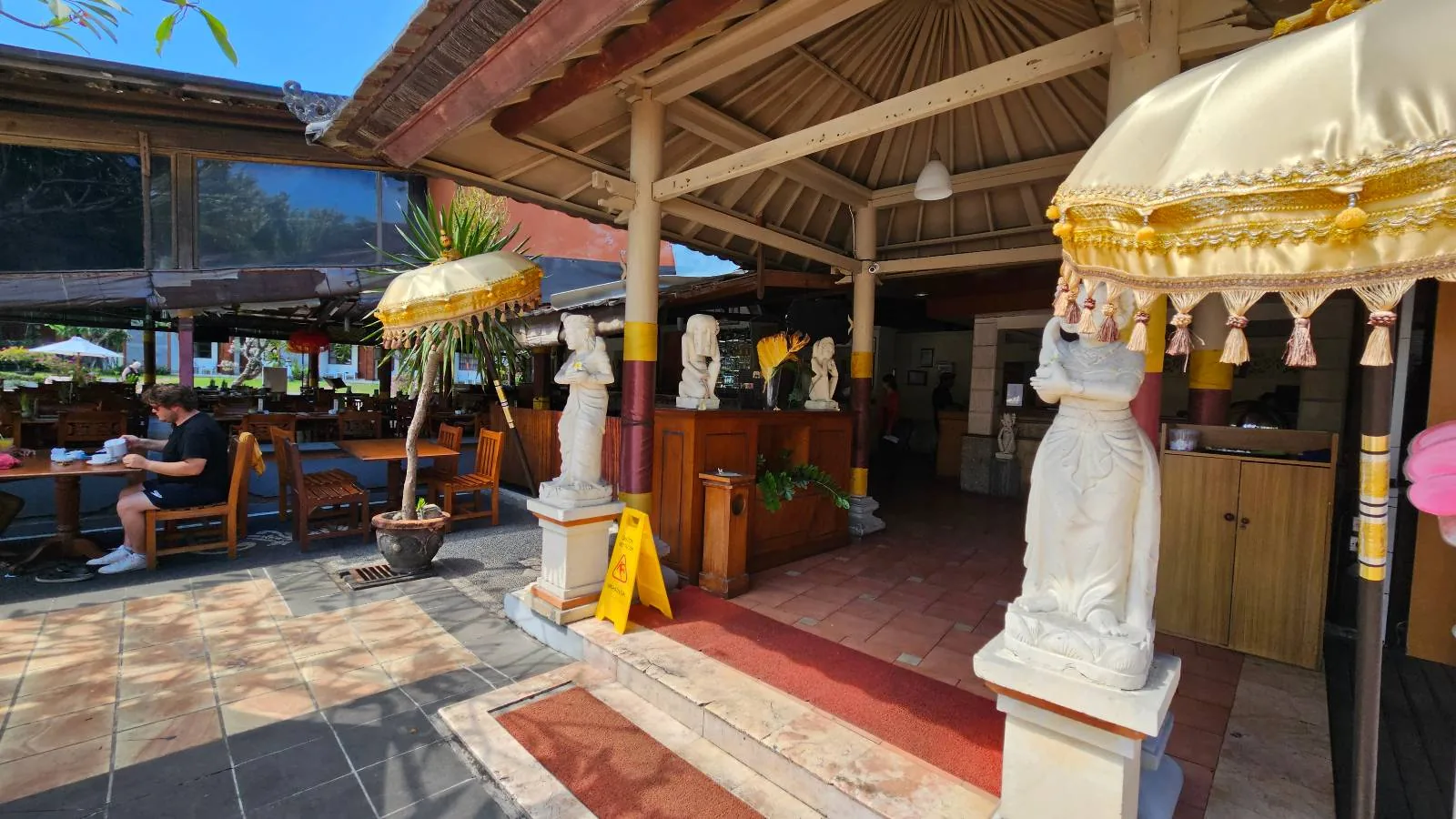 A tropical restaurant entrance with traditional statues and umbrellas. The interior has wooden structures and open seating. A wet floor sign is displayed. Lush greenery and outdoor seating are visible in the background.