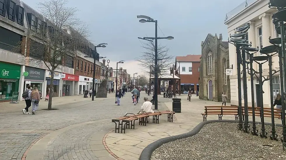 Pedestrians walk along a cobblestone street lined with shops and benches under a cloudy sky. A person sits on a bench. Urban setting.
