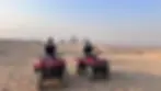 Two people on red ATVs in a desert with the Pyramids of Giza in the background. Clear sky and rocky terrain.