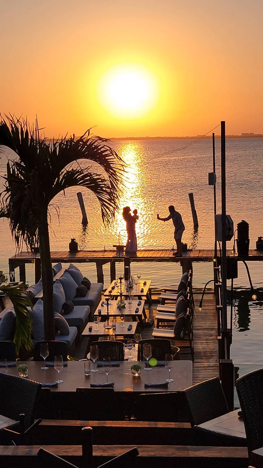 A silhouetted couple gently dance on a dock during a golden sunset over calm waters. Palm trees frame the scene, and a set table with chairs is visible in the foreground, adding a romantic atmosphere.