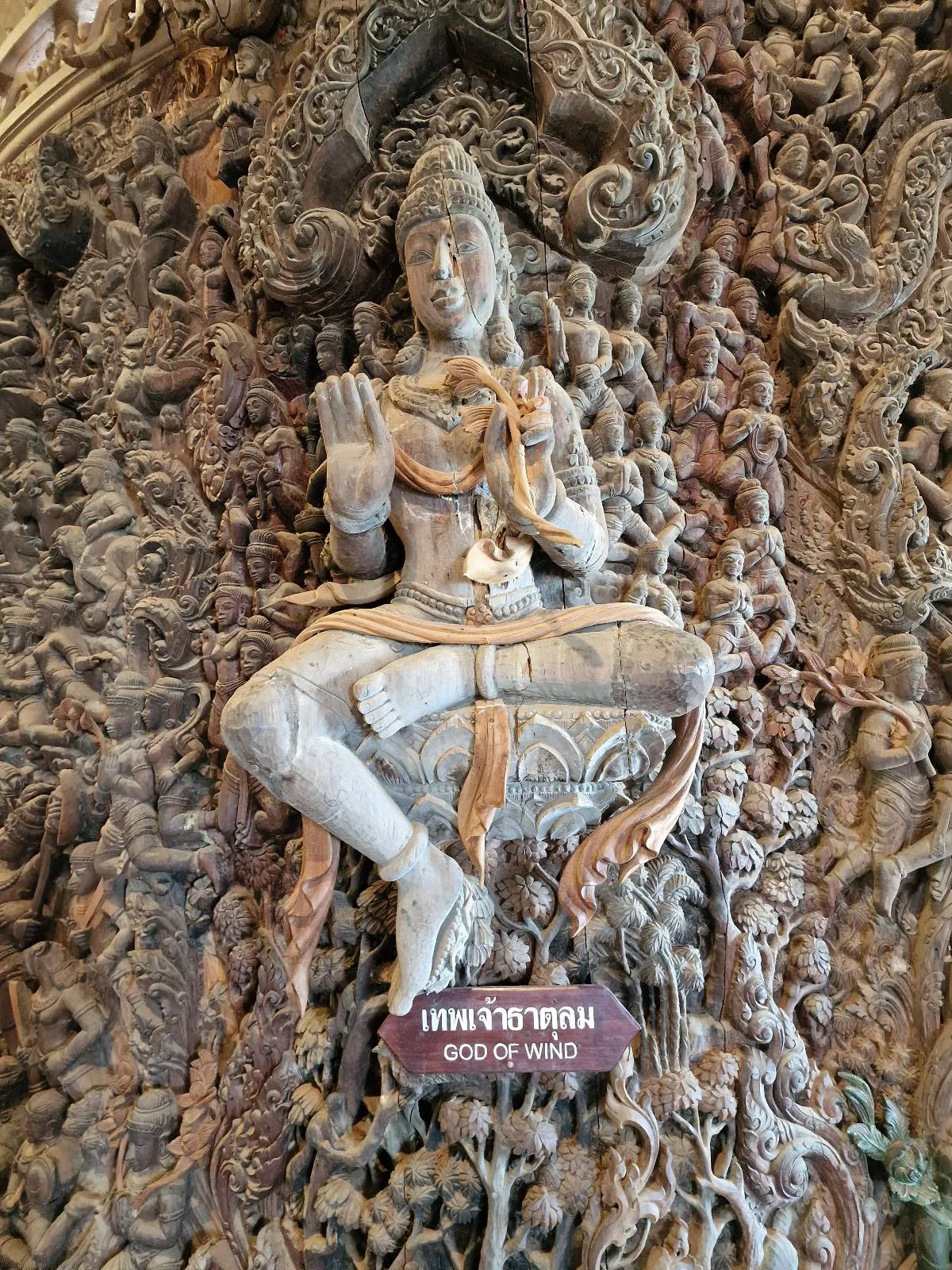 A detailed carving of a deity seated in a meditative pose, surrounded by intricately carved figures and patterns. A small sign below reads "GOD OF WIND." The art is highly detailed, showcasing craftsmanship and cultural significance.