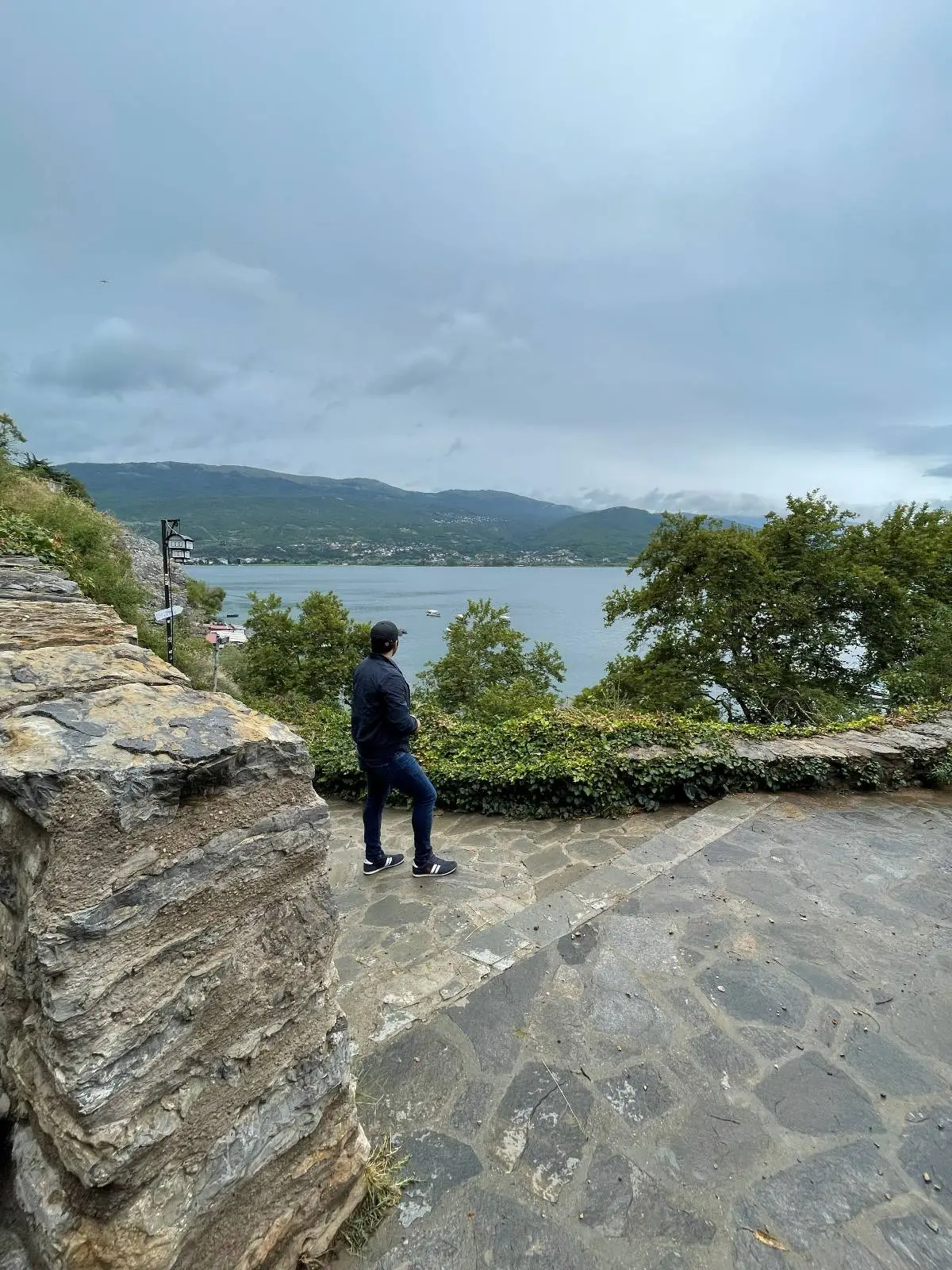 A person in a dark jacket and jeans stands on a stone pathway, looking out over a lake. The scene is surrounded by greenery, with mountains visible in the distance under a cloudy sky.