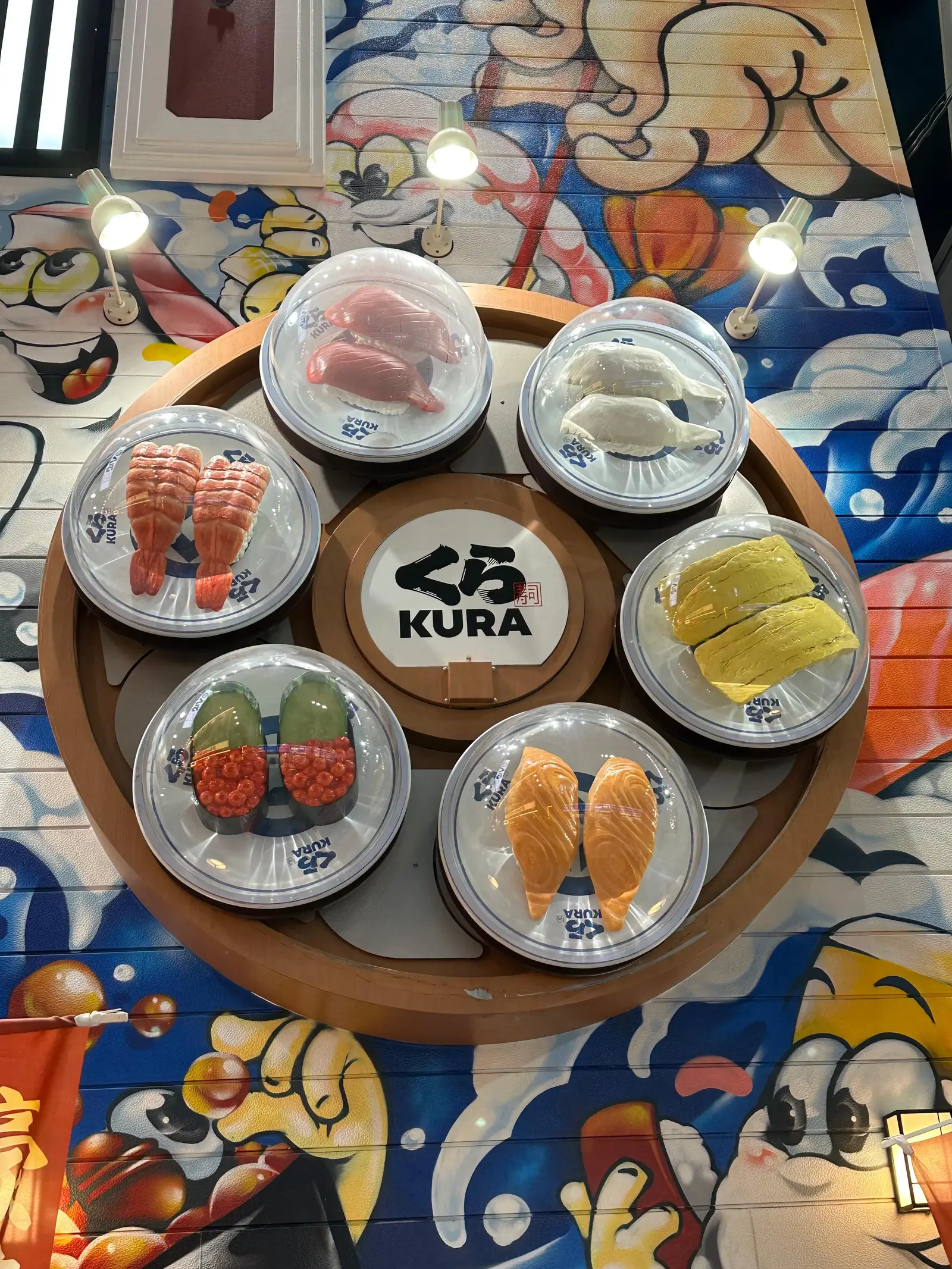 A round wooden tray displays six plates of assorted sushi around a central plate with the Kura logo. The tray is placed on a colorful, cartoon-themed table.