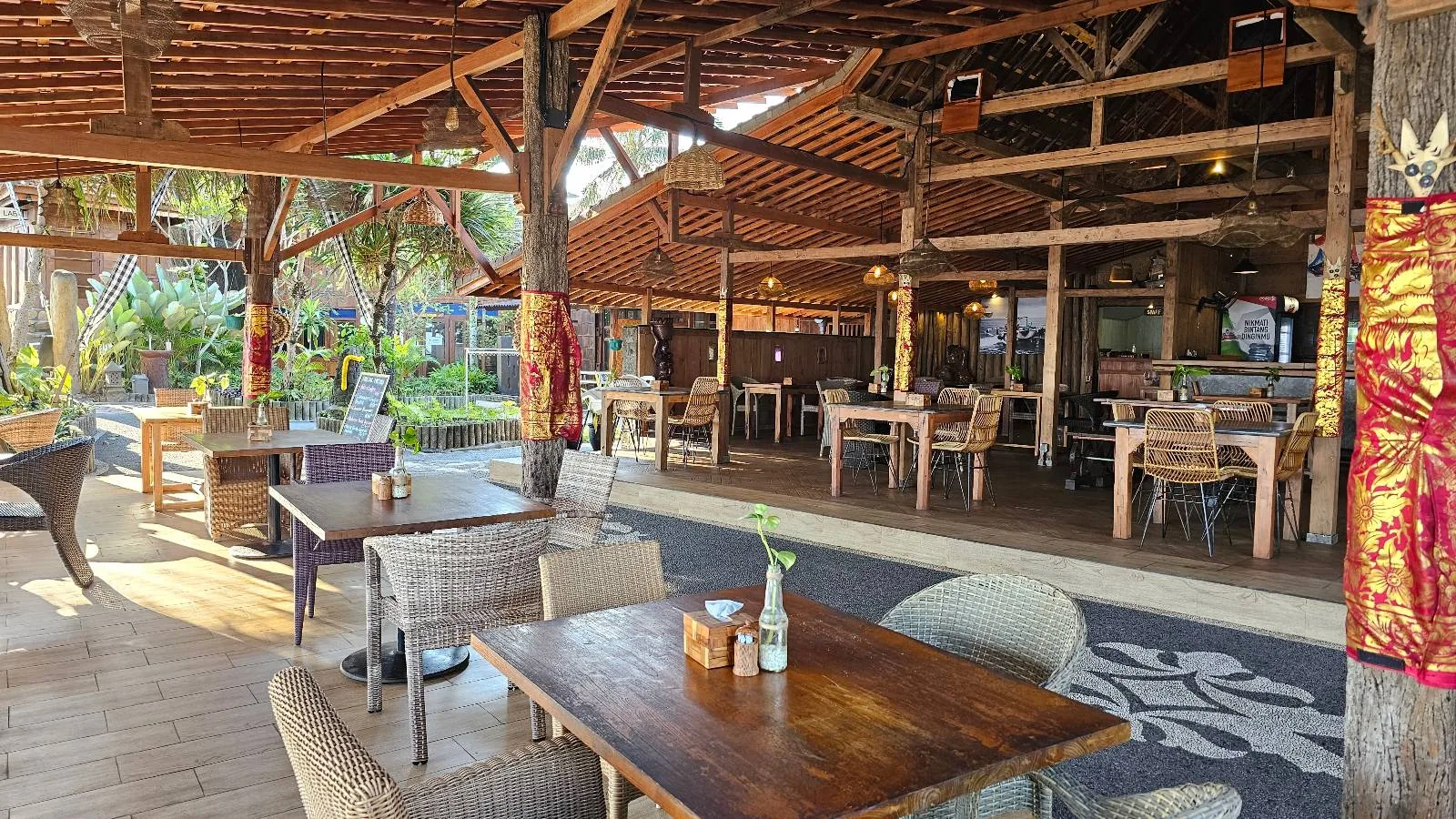 A person wearing a black outfit and cap sits at a wooden table on a seaside restaurant's patio. There's a small plant on the table. The ocean and beach are visible in the background with people walking along the shore.