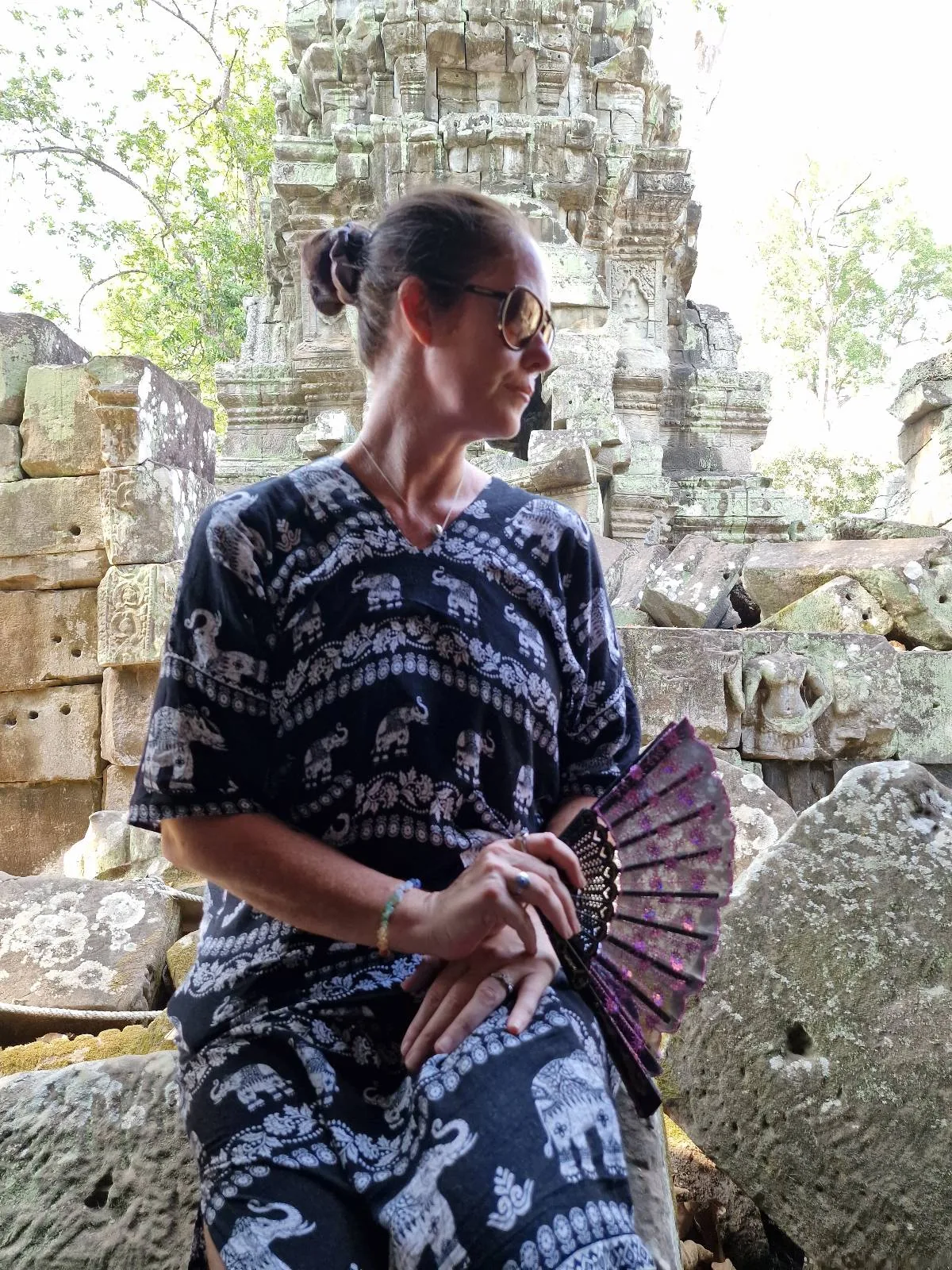 A woman wearing patterned clothing and holding a fan sits among ancient stone ruins, with weathered blocks and greenery visible in the background.