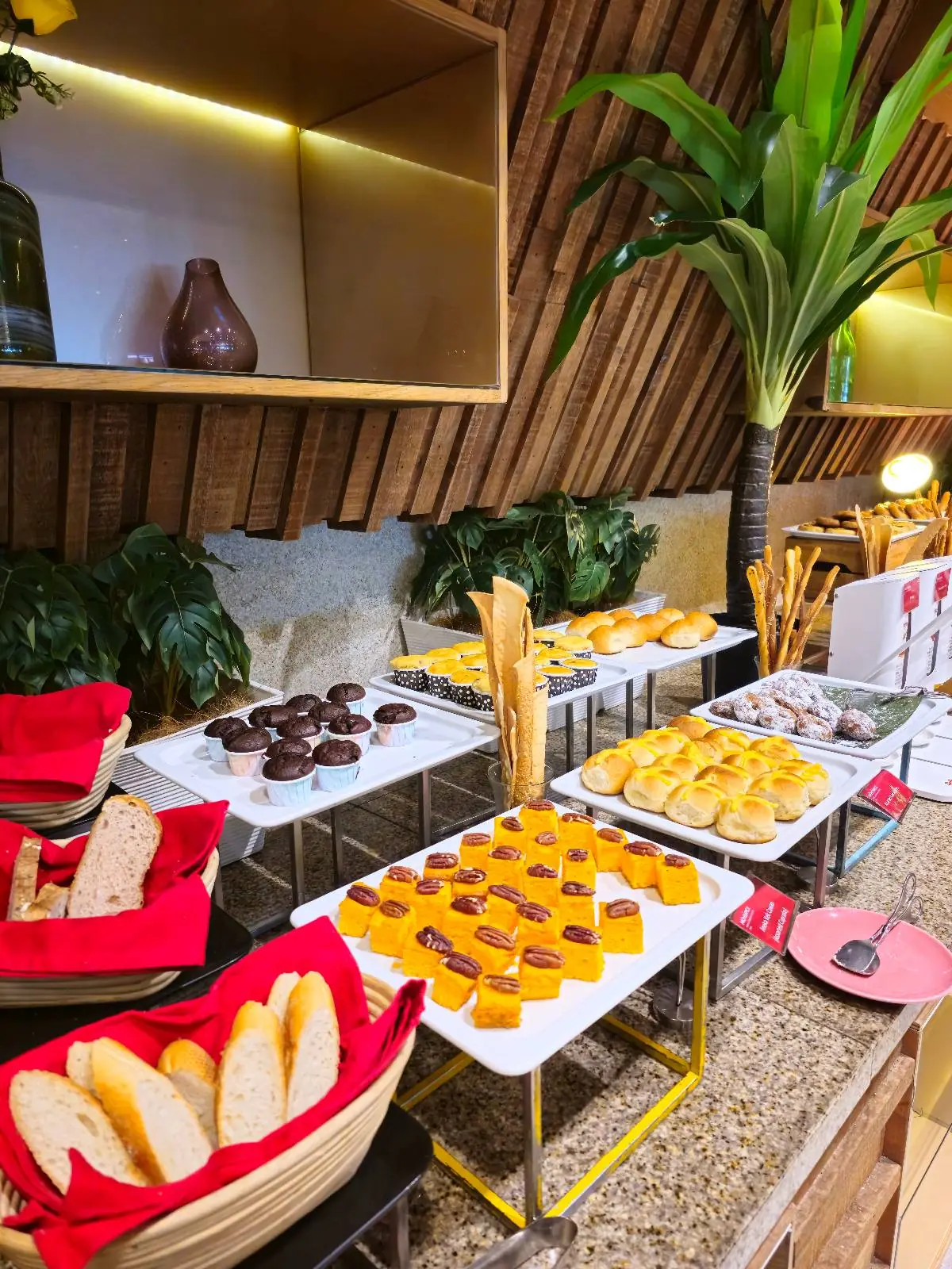A buffet table is adorned with a variety of pastries and breads, including baguettes in red baskets and neatly arranged desserts on white trays. The setting is enhanced by decorative plants and warm lighting, creating an inviting atmosphere.