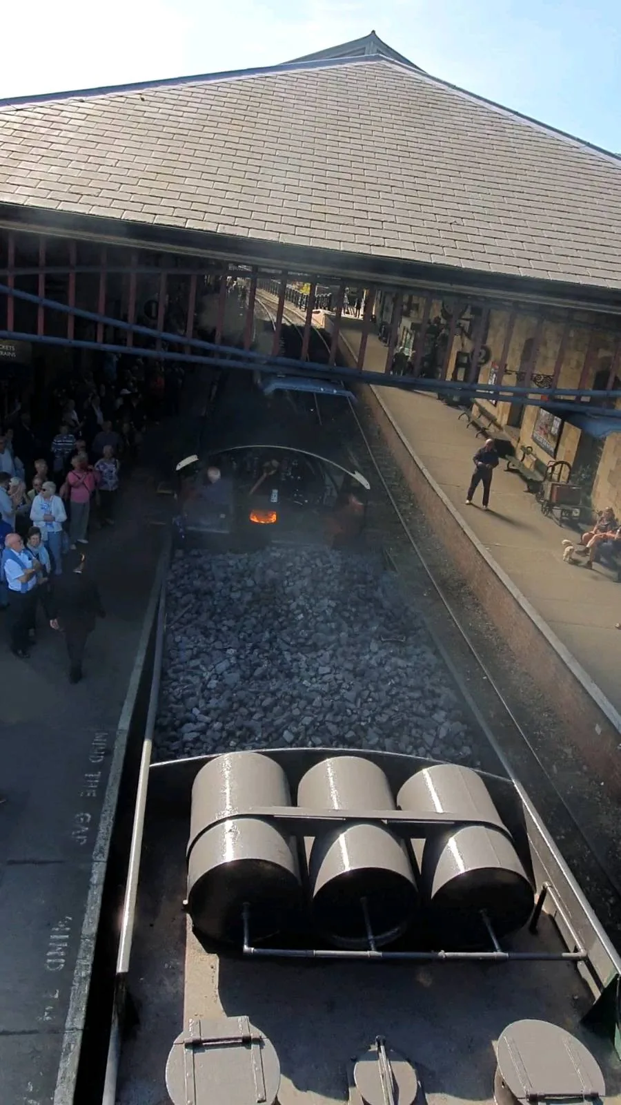 Aerial view of a historical railway station with people watching a heritage steam train. The train emits steam, and three barrels are visible on the platform. A large, roofed building surrounds the scene under a clear blue sky.