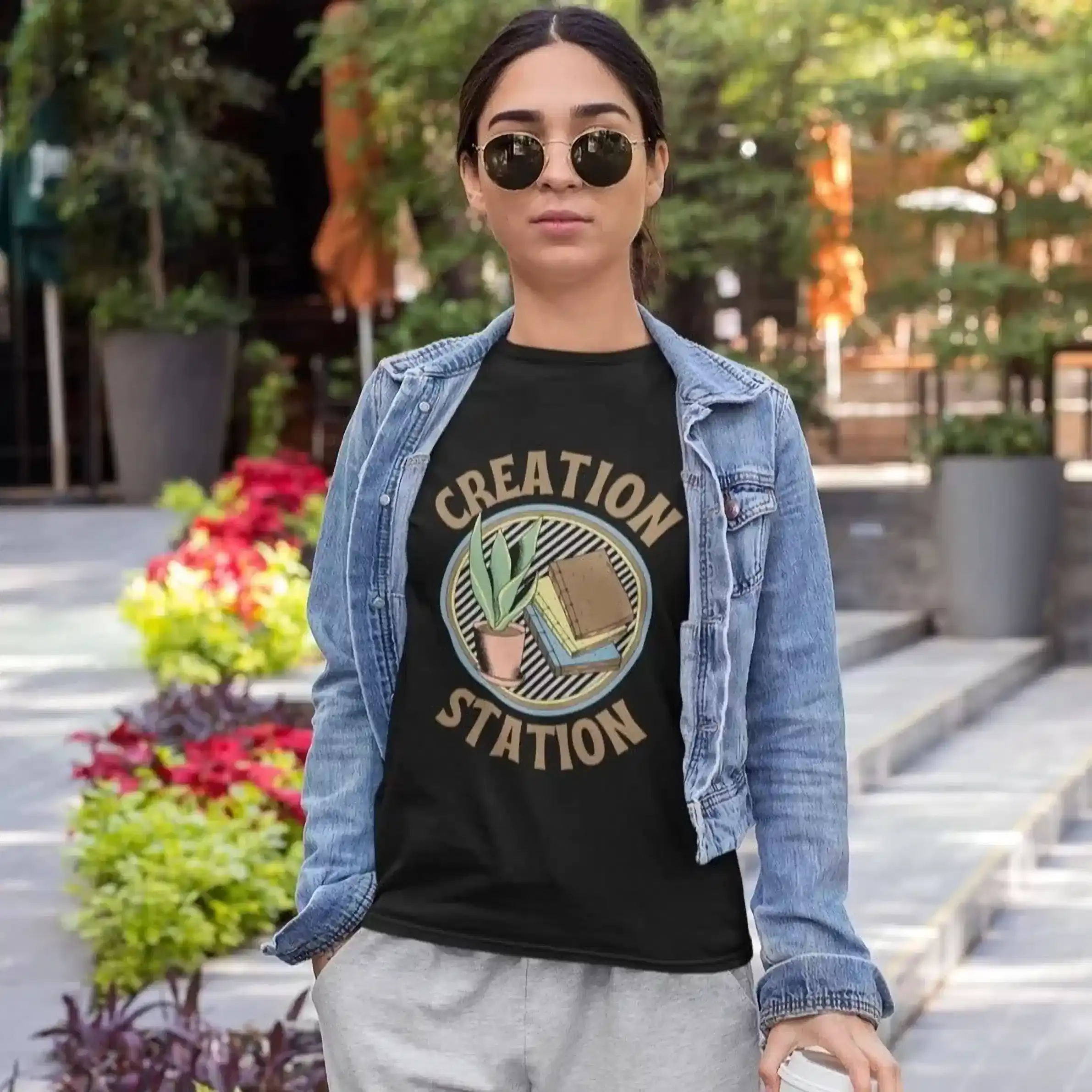 A person wearing sunglasses and a denim jacket walks down a city street, with a black t-shirt with "Creation Station" text.
