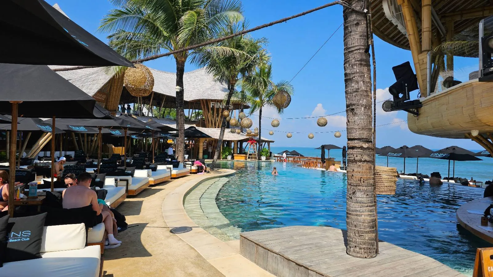 A tropical beachside bar with palm trees, a large pool with swim-up bar, and lounging guests under umbrellas. The ocean is visible in the background under a clear blue sky. Tables and seating line the pool's edge.