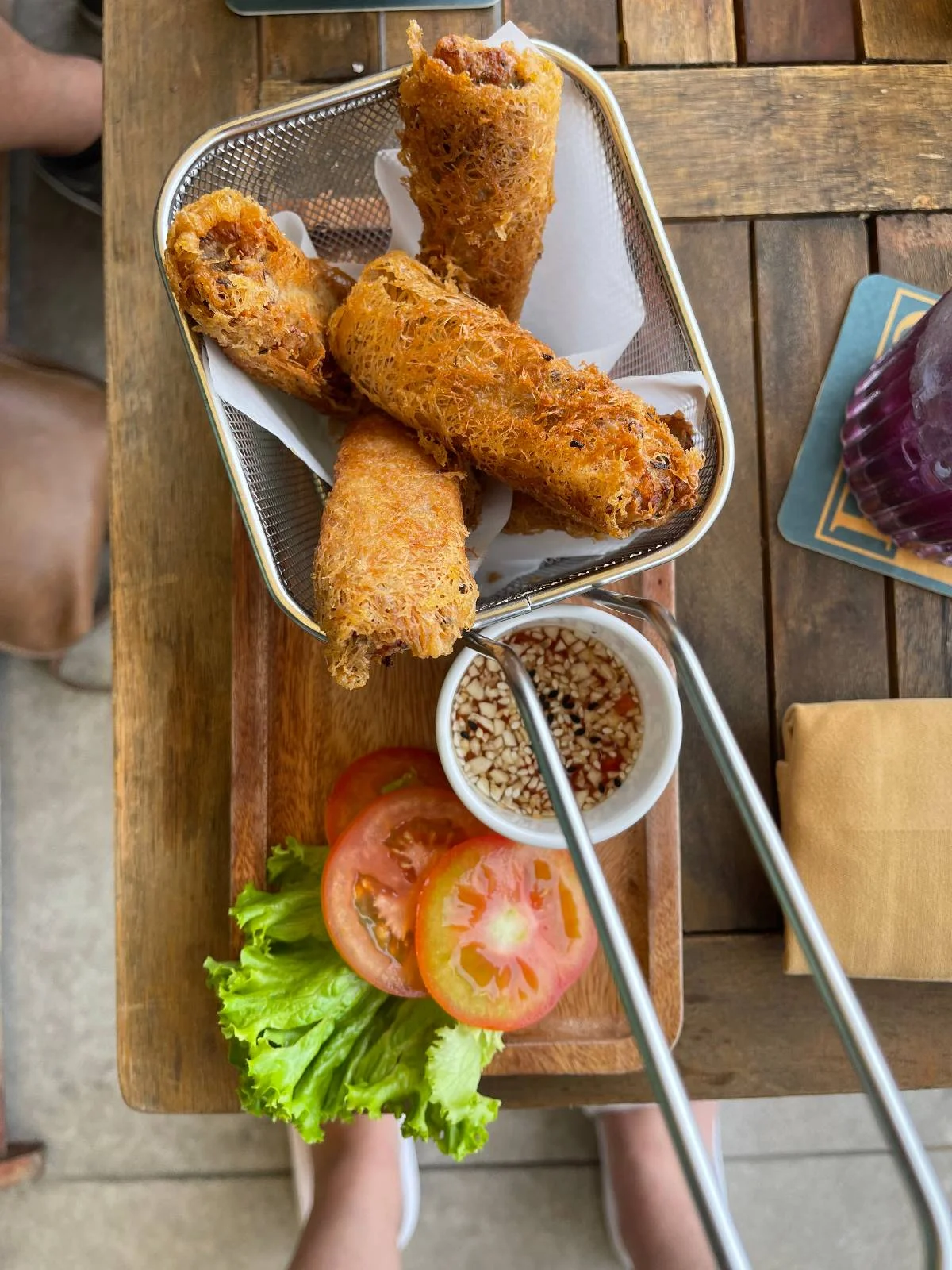 A small basket of fried spring rolls sits on a wooden table. Next to it are two slices of tomato and a few lettuce leaves. A dipping sauce with crushed nuts is beside the basket, along with a pair of chopsticks.