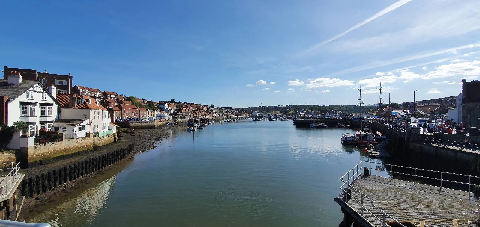 A tranquil harbor scene with calm water reflecting a clear blue sky. Historic buildings line the left bank, while boats and docks are visible on the right. A few fluffy clouds dot the horizon.