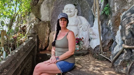 A woman seated beside a carved stone Buddha at Marble Mountain in Da Nang, surrounded by rock walls and greenery.