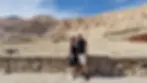 A couple poses in front of ancient cliffs and a temple under a clear blue sky. They are smiling, wearing casual clothes, next to a stone wall.