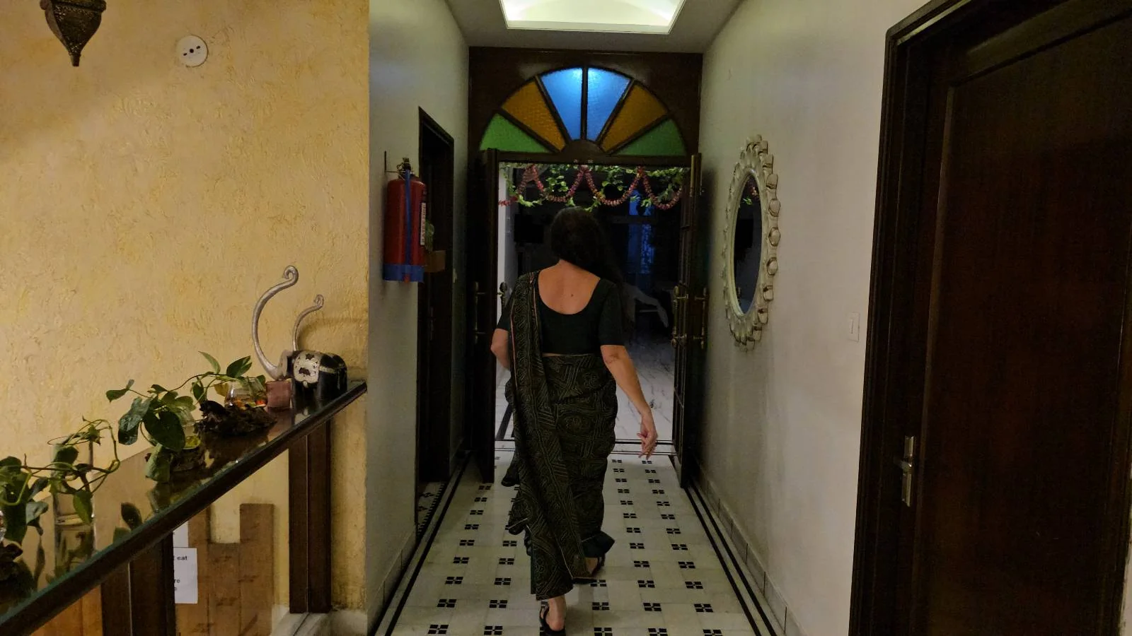 A person in dark clothing walks down a hallway with a tiled floor, plants on a side table, and colorful glass above a doorway at the end. The hallway is lit warmly and has a large round mirror on one wall.
