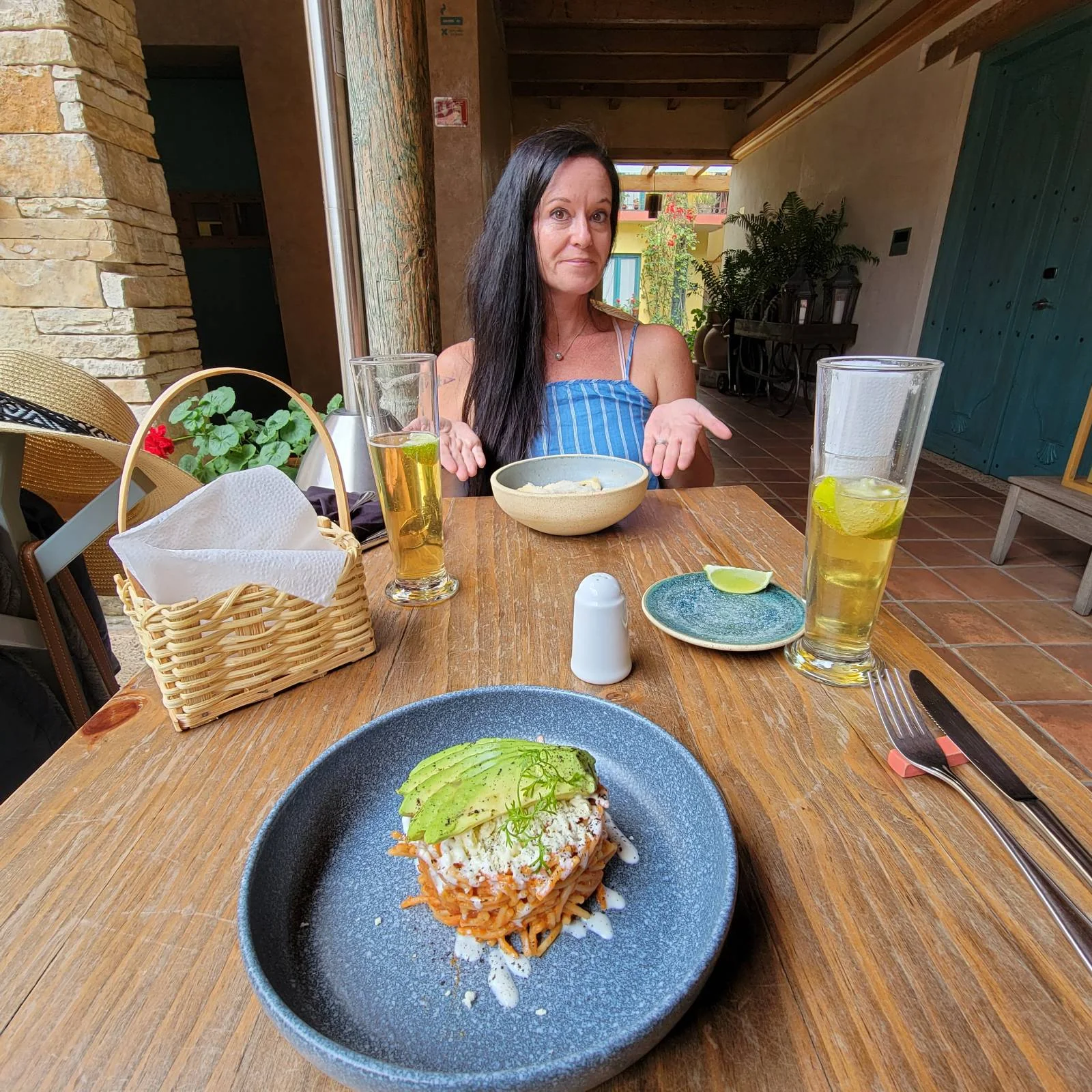 A woman with long dark hair sits at a table in a rustic restaurant. In front of her is a bowl and a tall drink with a lime. In the foreground, there's a plate with a dish topped with avocado slices.