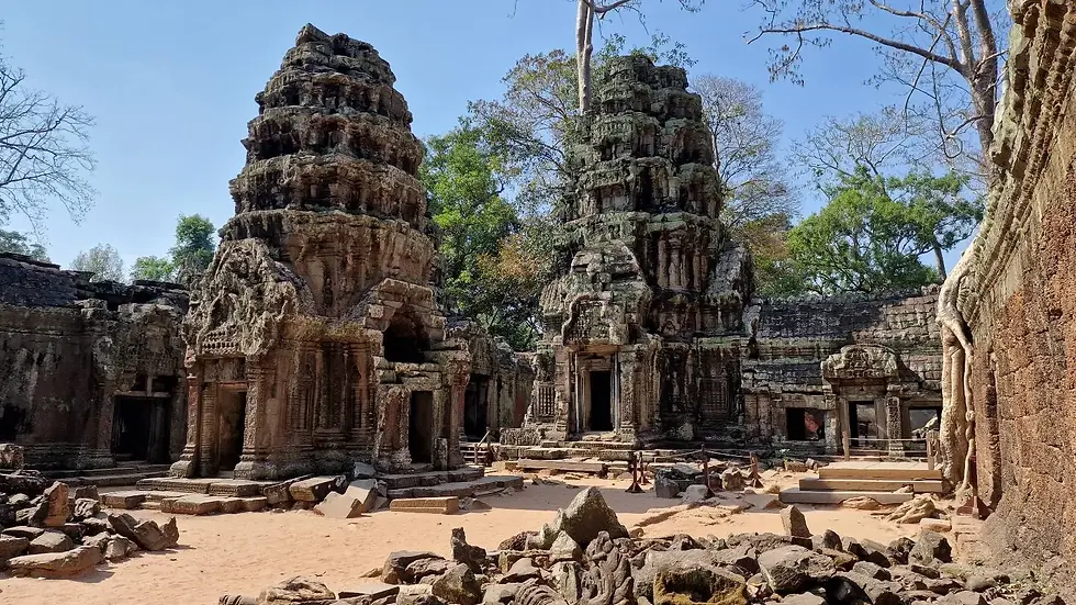 Ancient stone temple ruins with intricate carvings stand under a clear blue sky, surrounded by lush green trees. A serene, historic scene.