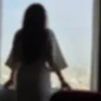 Silhouette of a woman in a robe looking out a large window over a cityscape. Her hands rest on the window ledge, capturing a reflective mood.