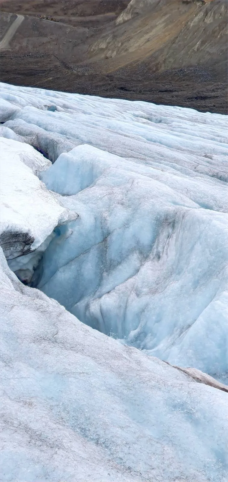 A close-up view of a glacier shows cracked, uneven ice with a deep crevice, set against a rocky, barren landscape in the background.