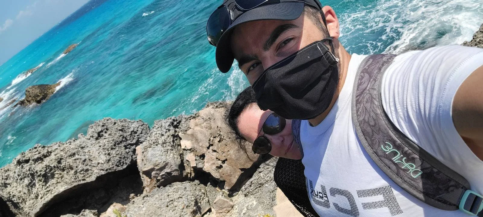 A person wearing a cap, sunglasses, and a mask takes a selfie with another person by a rocky shoreline. Behind them, the ocean is bright blue and clear on a sunny day.