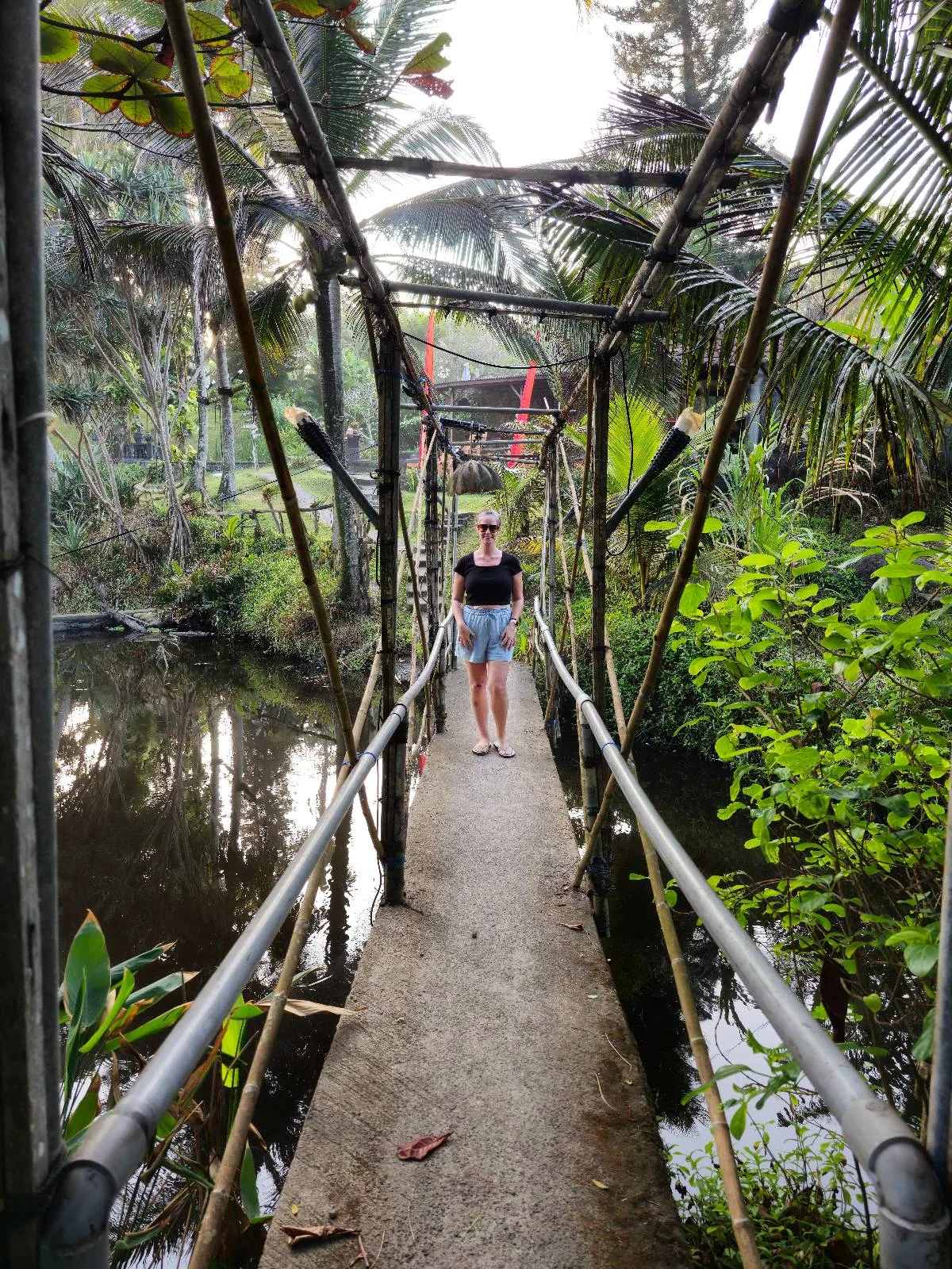 A person stands on a narrow, rustic bridge made of metal railings and concrete slabs, surrounded by lush green foliage and palm trees. The bridge crosses over a small body of water, creating a serene, natural setting.