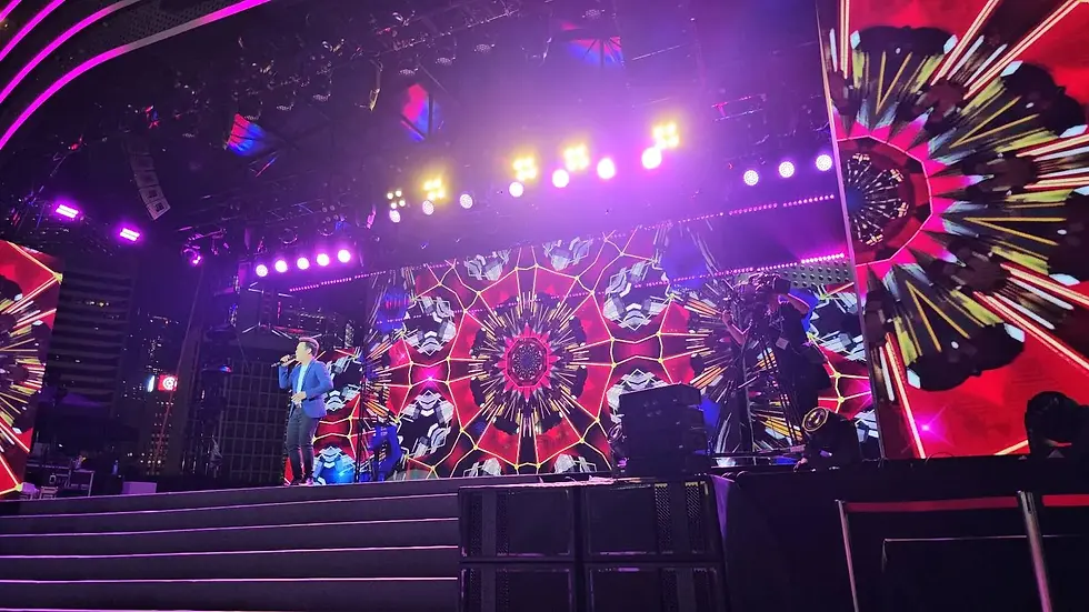 A person performs on a brightly lit stage with vibrant, kaleidoscopic patterns displayed on large screens in the background. The stage lighting is in shades of pink and purple.