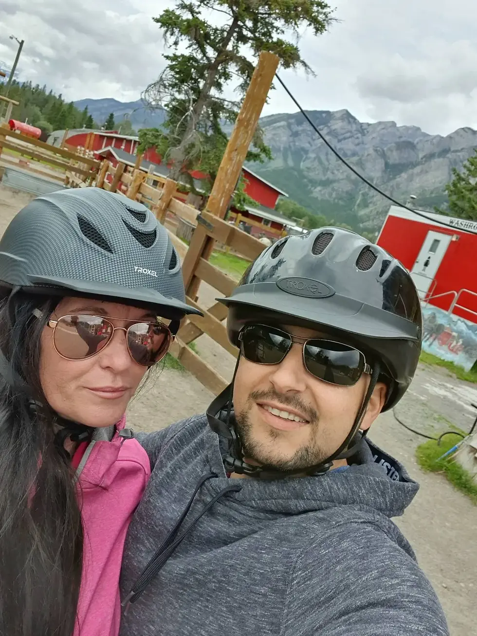 Two people wearing helmets and sunglasses pose for a selfie outdoors, with mountains and red buildings in the background.