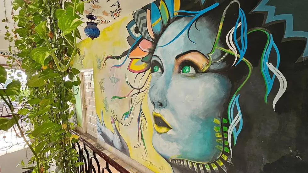 Mural of a face with vibrant colors and green eyes on a balcony wall. Lush green vines hang nearby, creating an artistic and serene setting.
