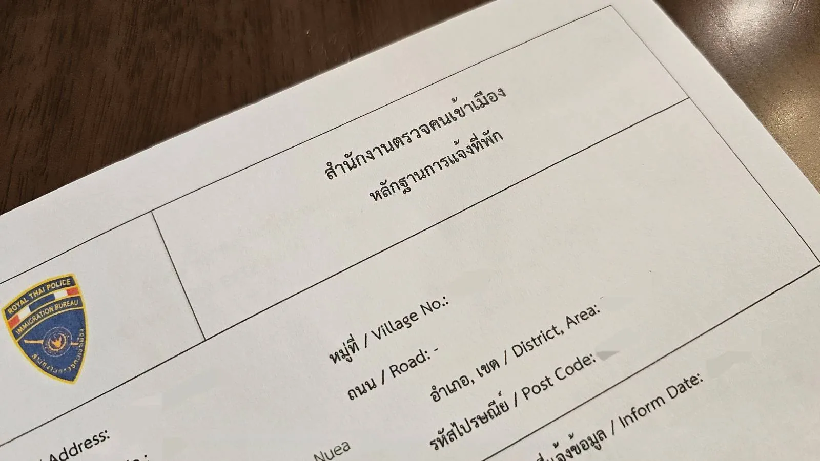 A close-up of a document with text in Thai and English. There is a crest or logo in the top left corner, and various fields for personal information such as name, address, and date of birth. The document rests on a brown surface.