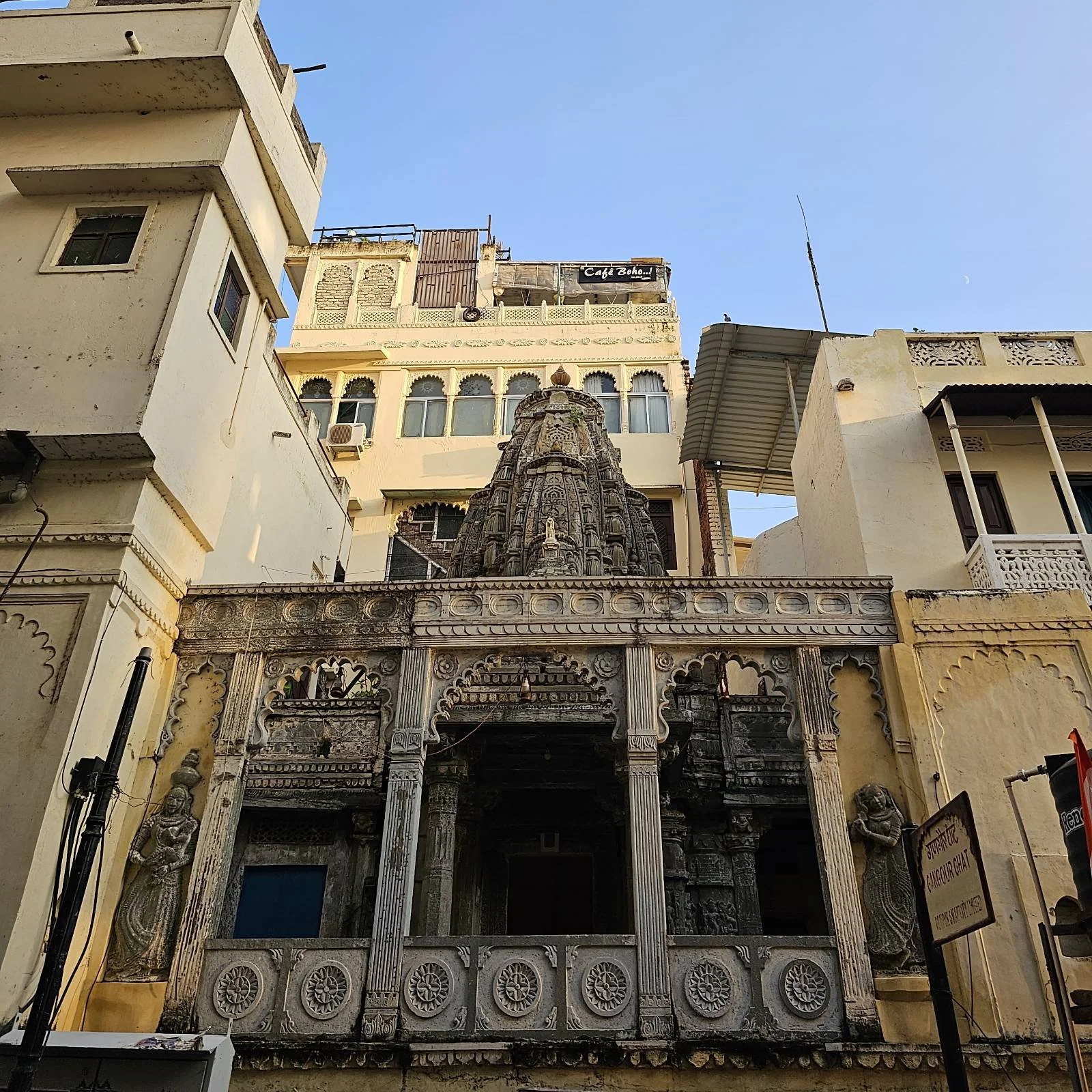 A stone temple with detailed carvings and pillars stands between taller, plain buildings under a clear blue sky. The temple entrance features ornate patterns and a small tower above it.