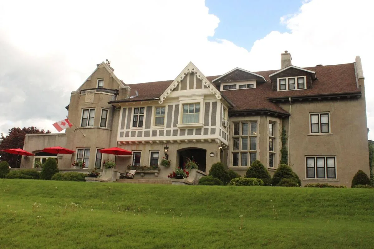 Historic stone building with red umbrellas, Canadian flag, lush green lawn, and cloudy sky, creating a serene and inviting atmosphere.