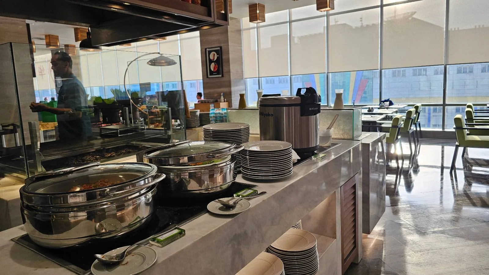 A bright, modern cafeteria with large windows, metal food warmers, stacked plates, and a rice cooker on a marble counter. A person stands behind the counter, and tables and chairs are visible in the background.