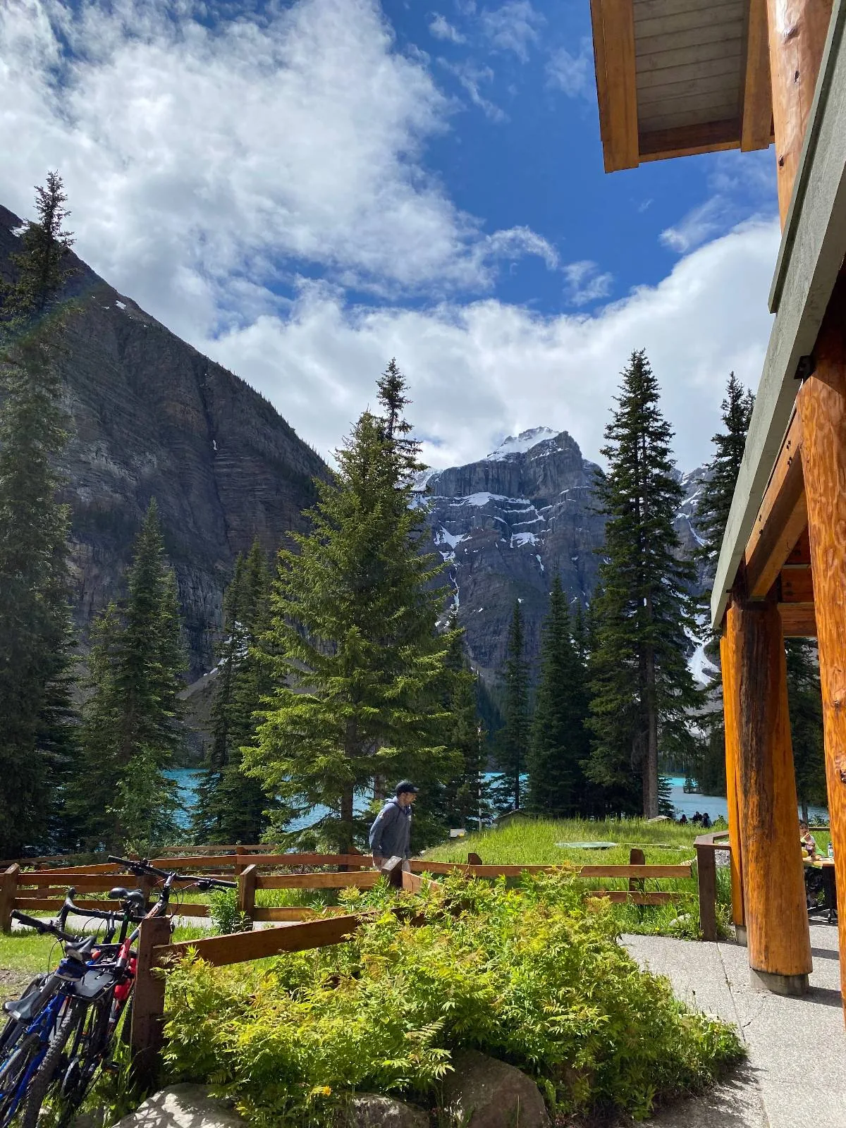 A scenic mountain landscape with trees, a clear blue sky with clouds, and snow-capped peaks in the background; bicycles are parked by a wooden fence and a wooden cabin is visible on the right.