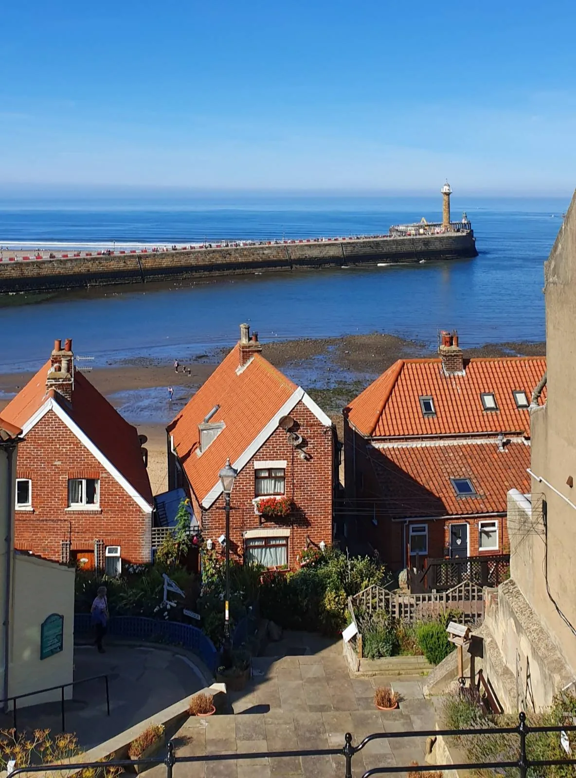 View of a coastal scene with a row of red-roofed houses in the foreground. A pier extends into the blue sea, topped with a lighthouse. The sky is clear and bright, highlighting the calm, scenic atmosphere.