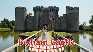 Woman in a blue dress walks on a bridge towards Bodiam Castle over a moat, with "Bodiam Castle" text at the bottom with red and white flags.