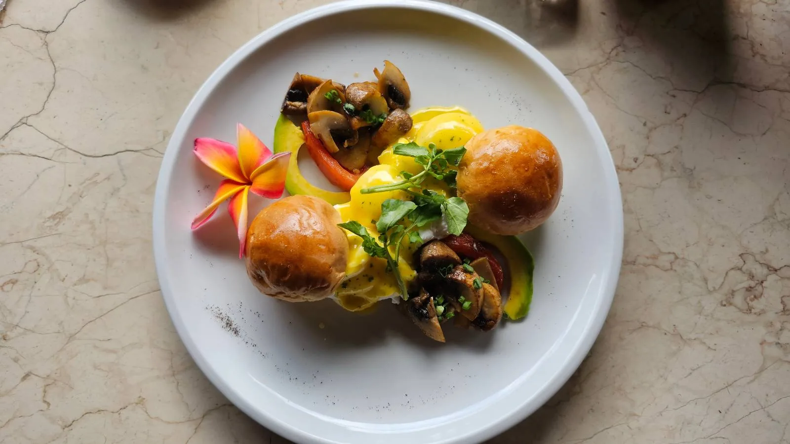A white plate with two golden buns, topped with creamy sauce, garnished with sautéed mushrooms, greens, and a small pink and yellow flower. The arrangement is artistic and vibrant, set on a marble table.