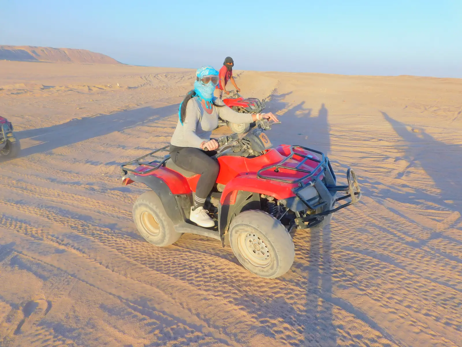 A person rides an ATV on a sandy landscape under a clear blue sky. Two hills are visible in the background, along with a small building structure. The scene conveys a sense of adventure in a desert setting.