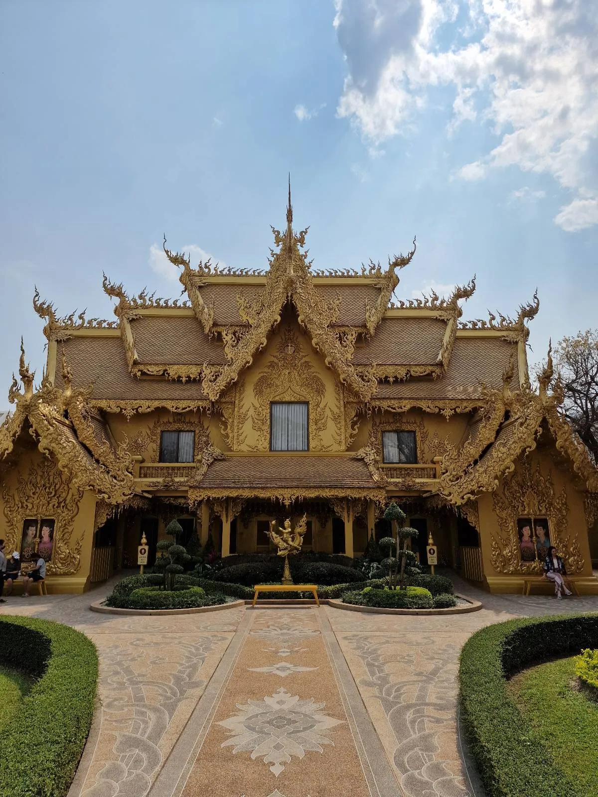 A golden temple with intricate architectural details under a partly cloudy sky. The temple features ornate spires and decorative carvings. A symmetrical garden with manicured bushes leads up to the entrance of the building.