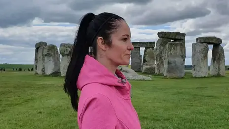 Woman standing near the Stonehenge stone circle in Wiltshire, England, with ancient standing stones set across open grassland under a cloudy sky.