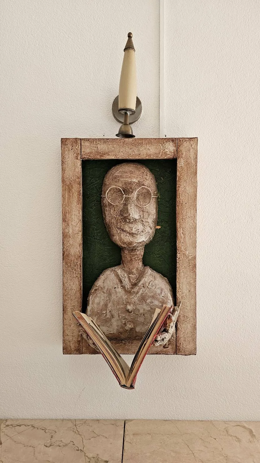 A framed sculpture of a human bust with a rough texture is mounted on a wall; an open book is placed below the frame, and a candle holder sits on top of the frame.