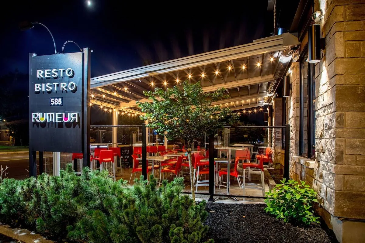 Outdoor view of a well-lit Resto Bistro patio at night, featuring red chairs, tables, and string lights under a pergola. A large sign displays the restaurant's name and street number. Greenery surrounds the patio area.