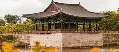 A traditional Korean wooden pavilion with a tiered tiled roof, sitting on a high stone platform surrounded by a reflective pond and lush trees.