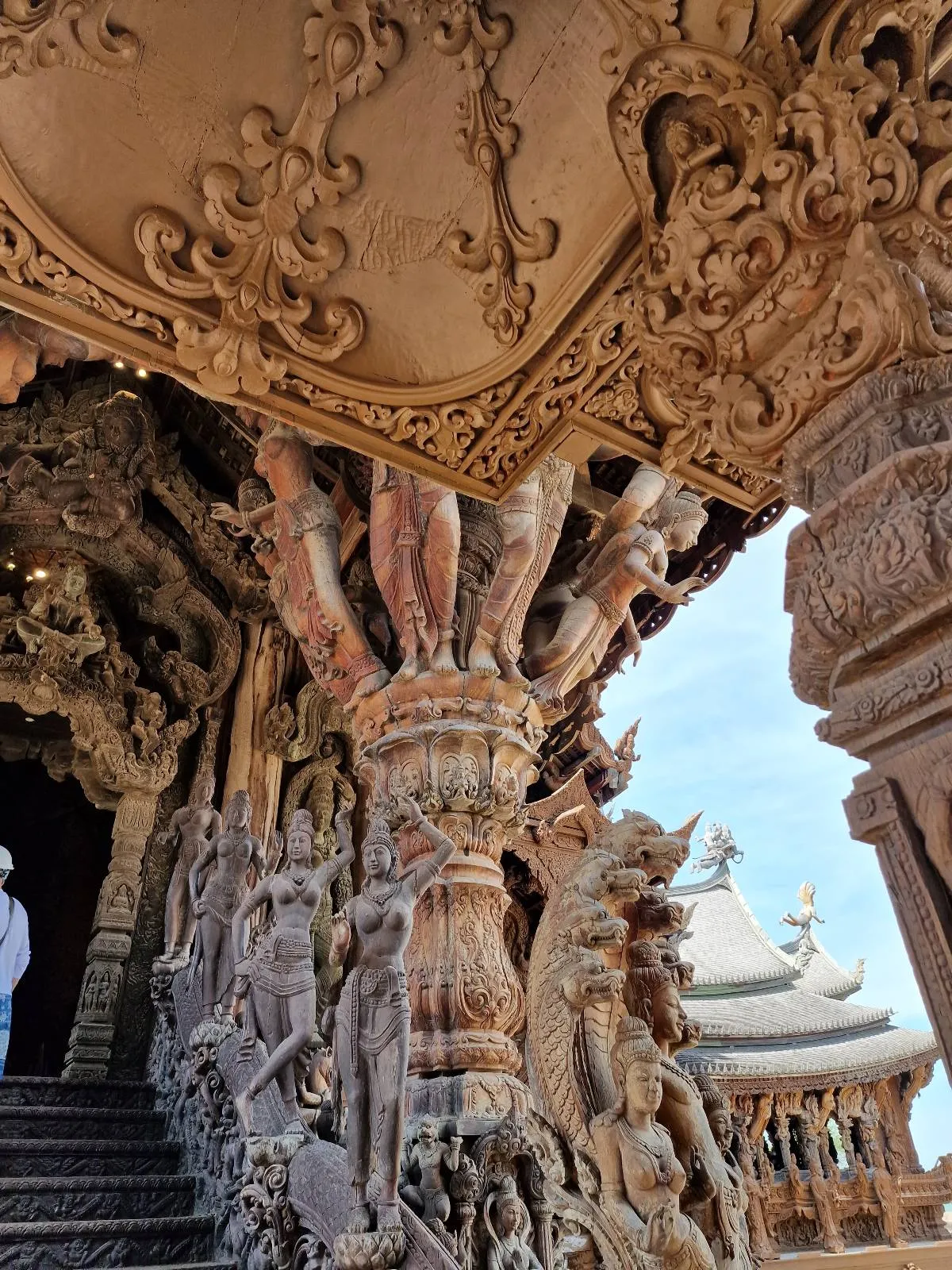 Intricately carved wooden columns and sculptures depicting mythical figures are seen at the entrance of an elaborate temple. The detailed craftsmanship is highlighted against a clear blue sky.