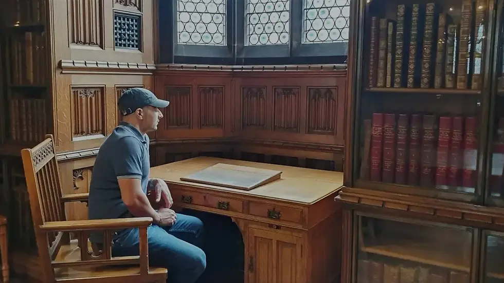 Man in cap sits at a wooden desk in a library with ornate wood paneling and bookshelves. The setting feels historic and reflective.