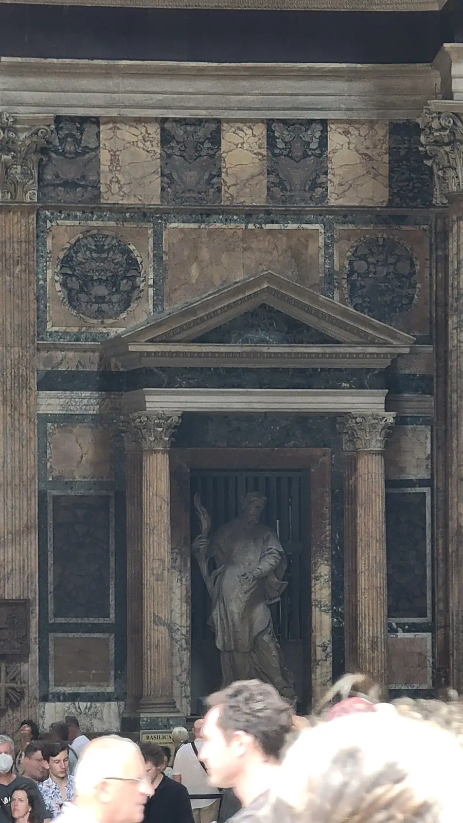 A stone statue stands in a dark alcove framed by columns and an ornate rectangular archway. The façade features patterned stonework, decorative carvings, and reliefs, with several people visible at the bottom of the image.