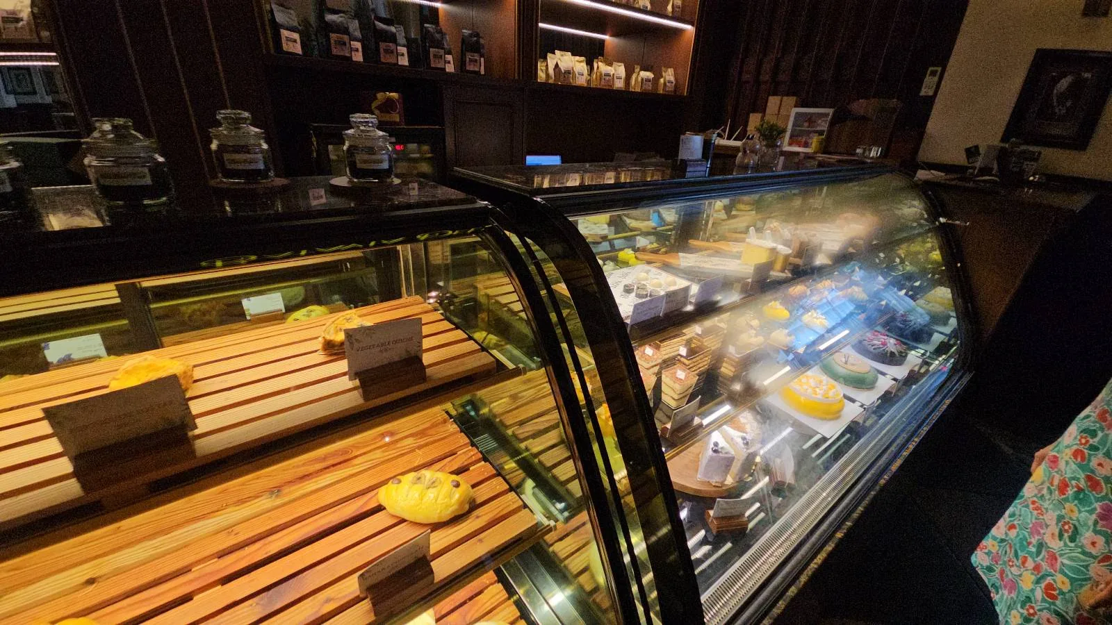 A pastry display case in a dimly lit bakery. The case contains assorted pastries, breads, and desserts on wooden shelves. Behind the case, shelves with jars and decor are visible. The atmosphere is cozy and inviting.