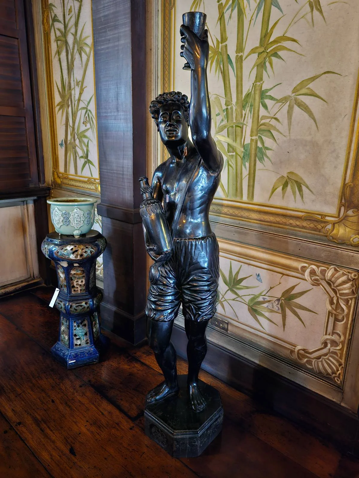 A bronze statue of a man in shorts holds a torch aloft. He stands on a pedestal with one arm raised. Next to him, a decorative vase sits on a stand. The backdrop features a bamboo-patterned wall.