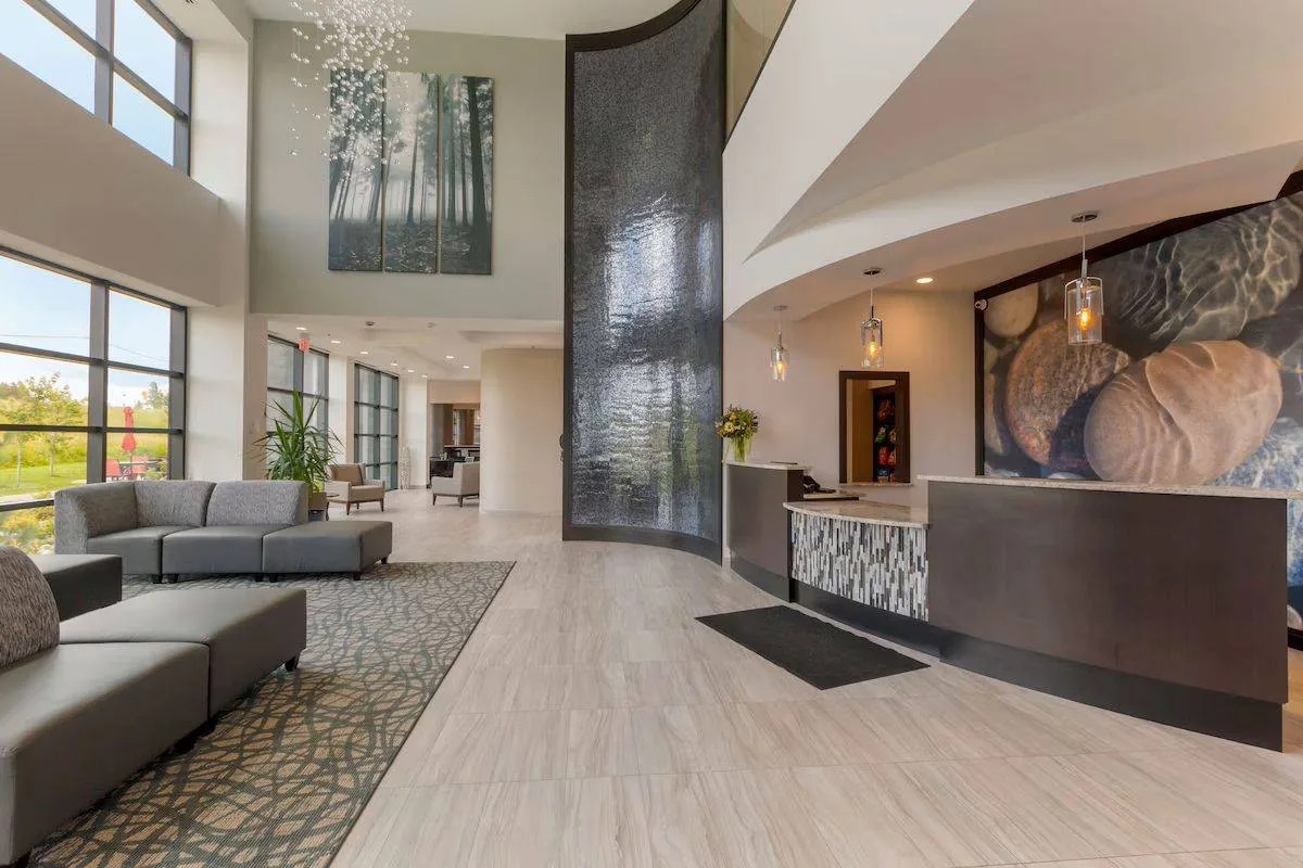 Modern lobby with polished tile floors, a curved reception desk, and a stone accent wall featuring shell artwork. Large windows on the left let in natural light, and there are gray sofas on a patterned rug. A tall, abstract art piece hangs above.