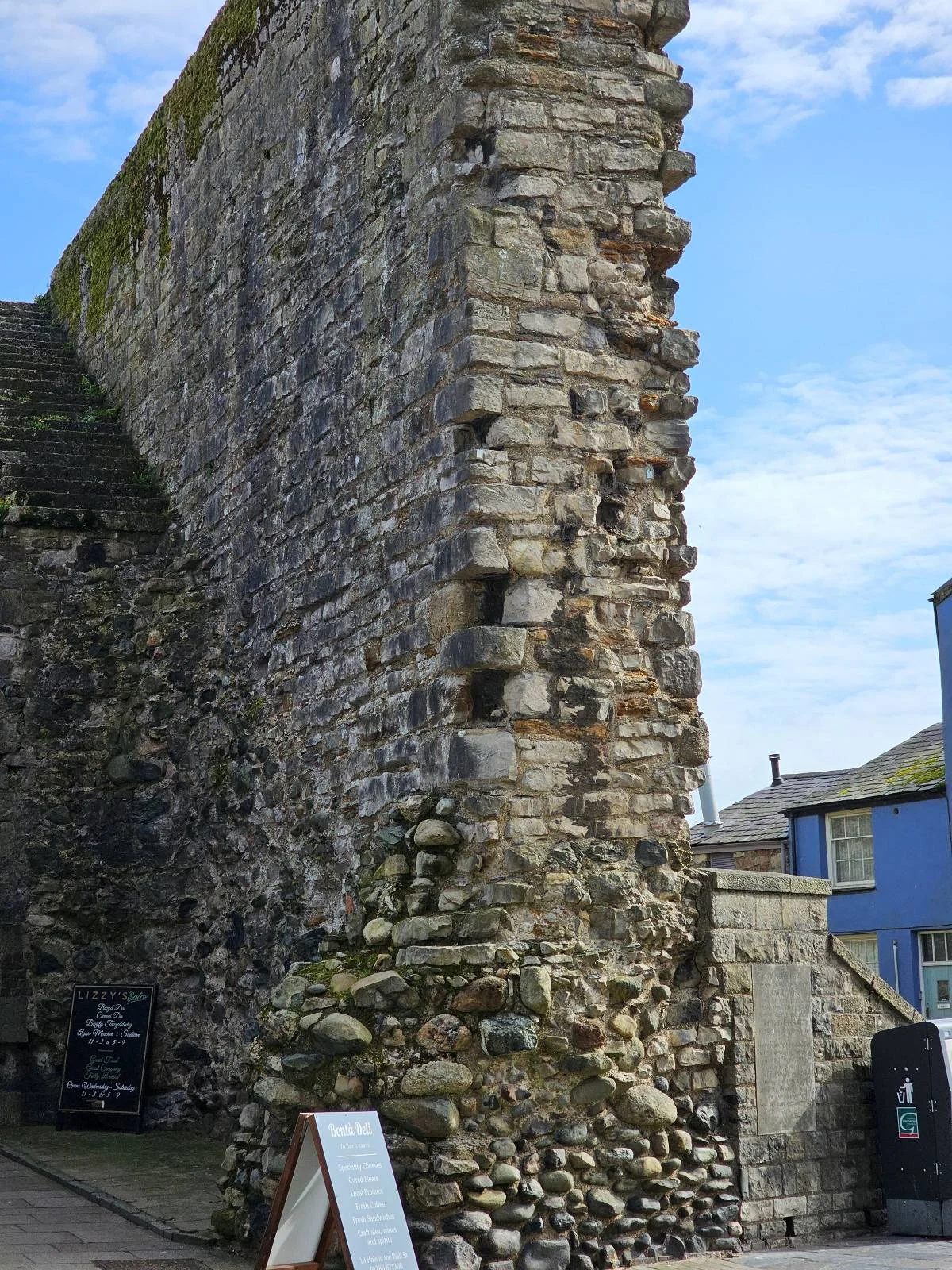 A tall, weathered stone wall stands outdoors, showing signs of age and crumbling in some areas. In the background, there are modern buildings, including a blue house, under a partly cloudy sky.