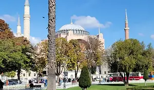 Hagia Sophia in Istanbul seen from a park with trees and crowds, its central dome and minarets set against a clear blue sky.